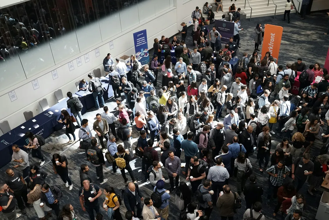 A conference expo floor seen from the mezzanine: hundreds of attendees with lanyards, phones and laptop bags packed between sponsor tables